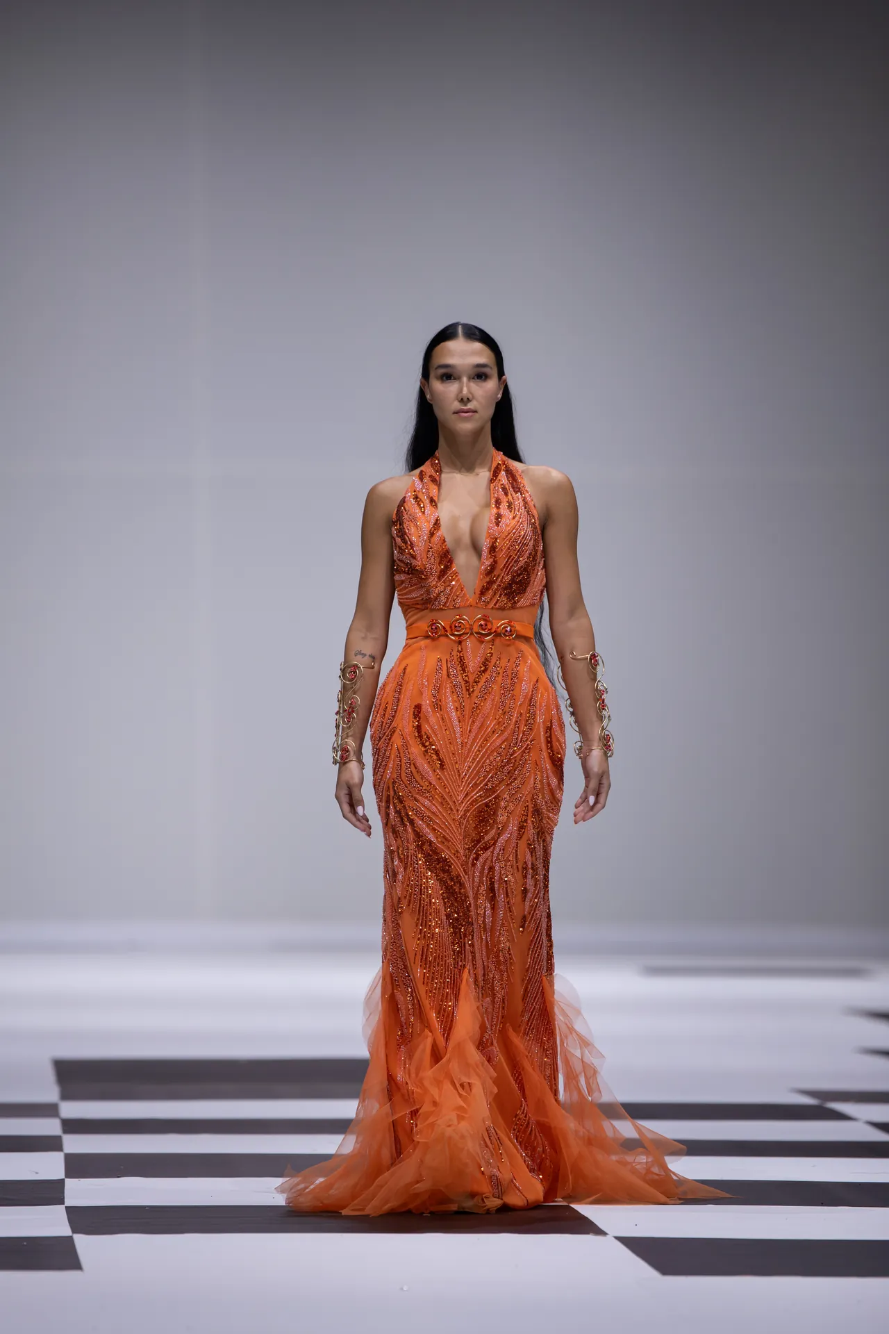 Model walks the runway in a bright orange sequined halter gown with a plunging neckline and a sheer train.