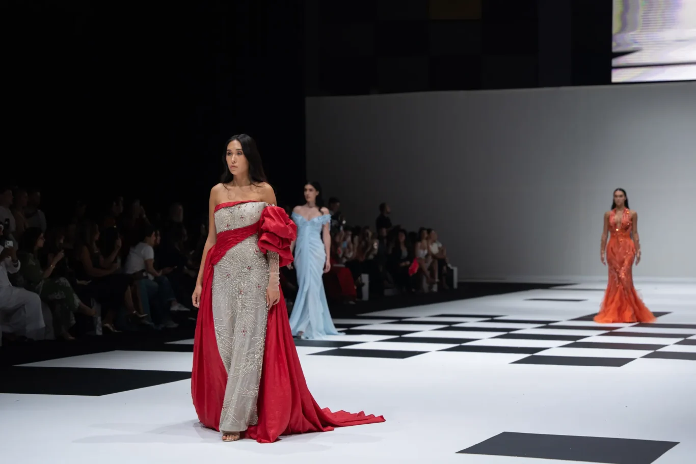 Model walks a fashion runway in a sparkling silver gown with a red cape, on a black-and-white checkered catwalk, with other models and audience in the background.