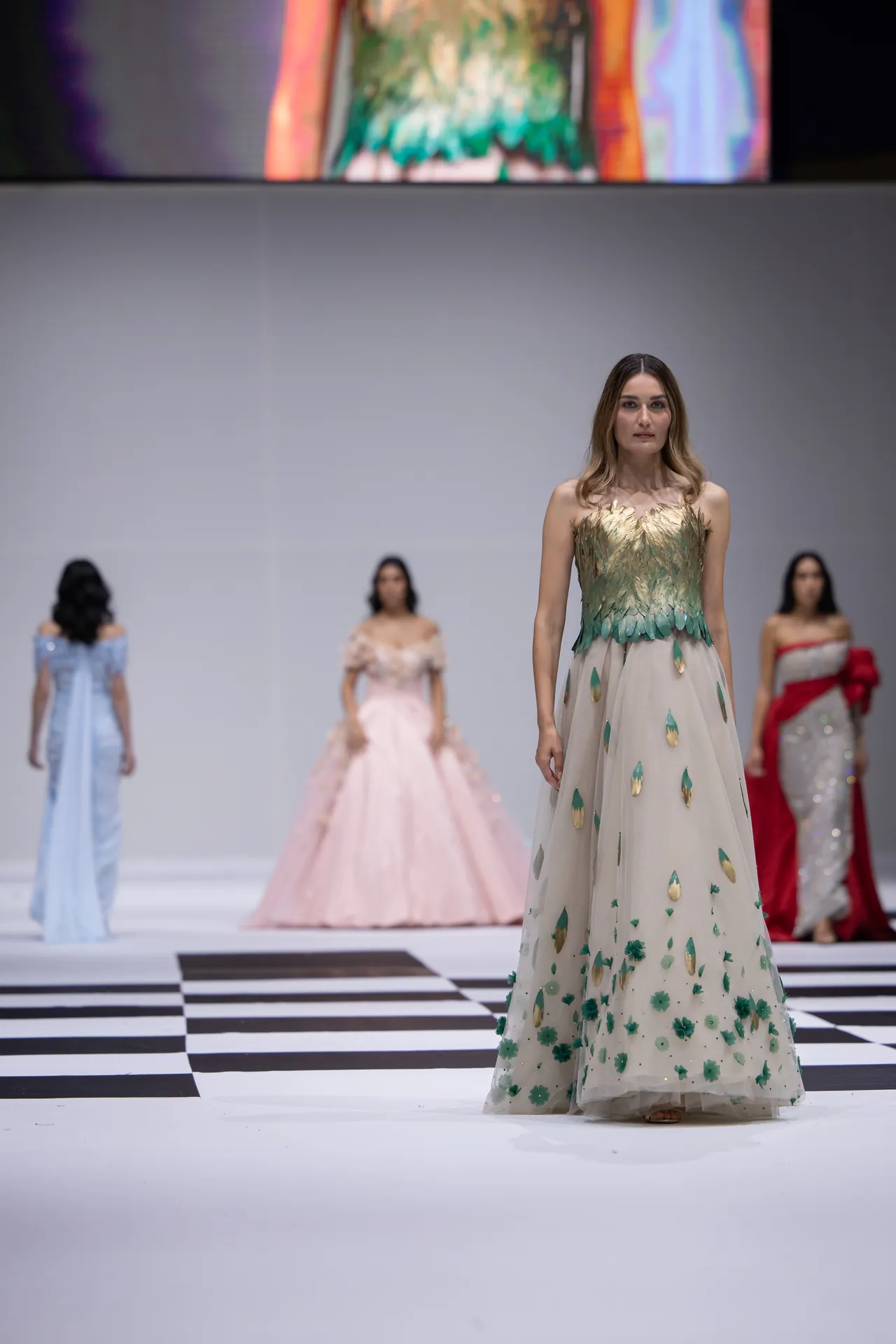 Model on a fashion runway wears a pale gown with green leaf-like embellishments; checkered floor and blurred background models visible.