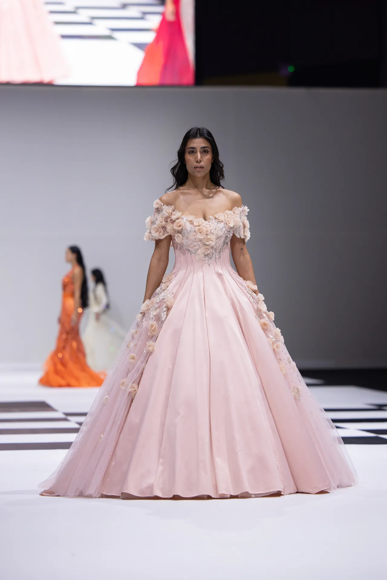 Model on a runway wearing a pale pink off-the-shoulder ball gown with floral appliqués walking the catwalk.