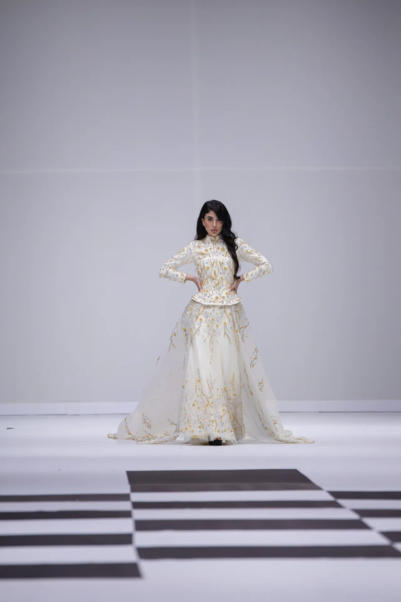 Female model in an ornate white gown with gold embroidery standing on a minimalist runway.