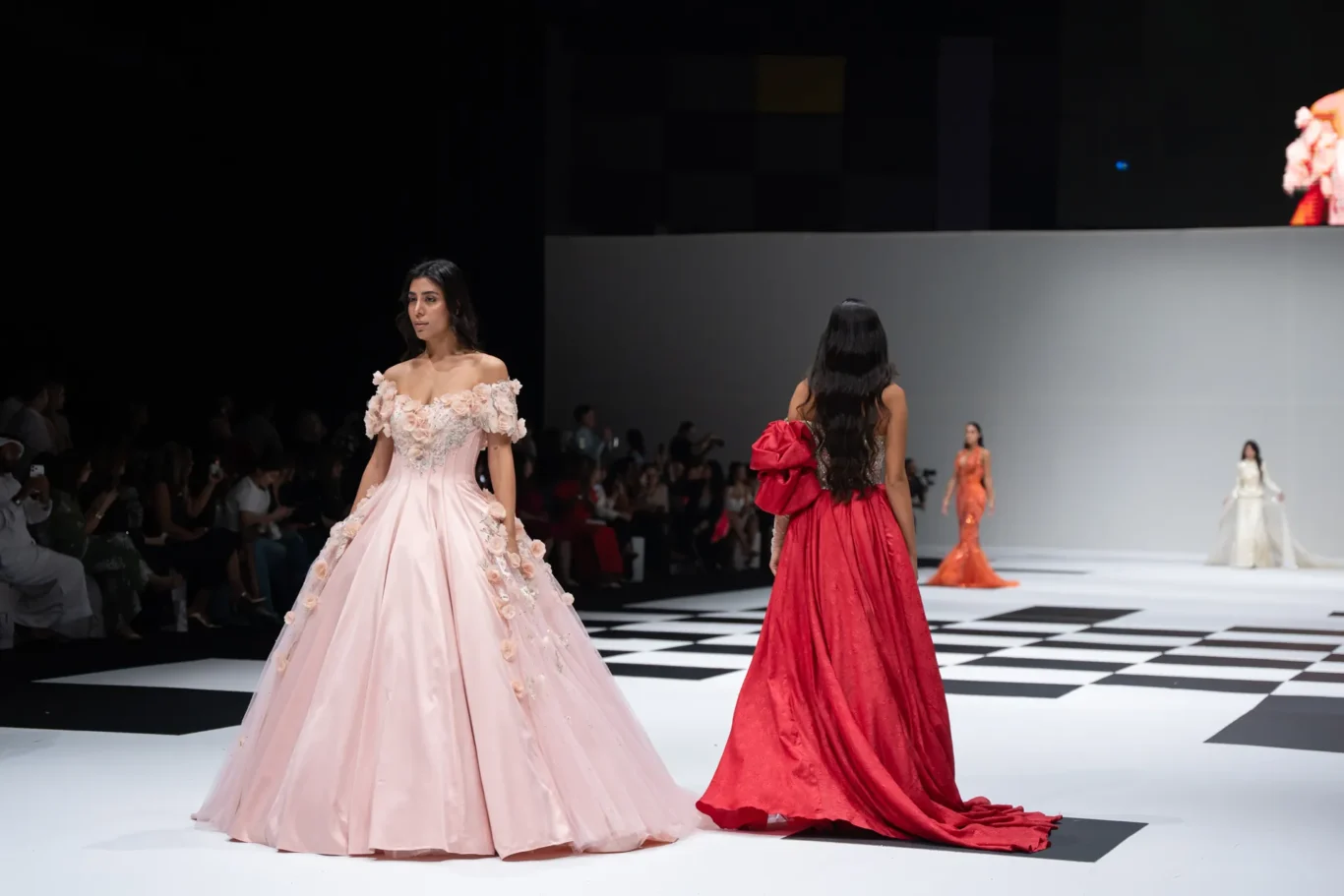 Models walk a checkerboard runway in elegant gowns; a pale pink off-shoulder ball gown with floral appliqués leads, while a red gown trails behind.