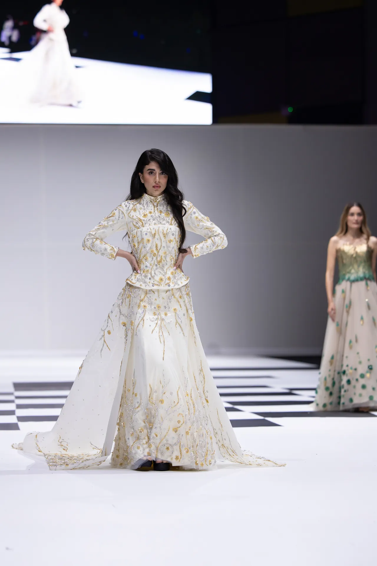Model on a runway wearing a white, gold-embroidered gown with hands on hips, checkerboard floor in foreground, other model in a green dress in background.