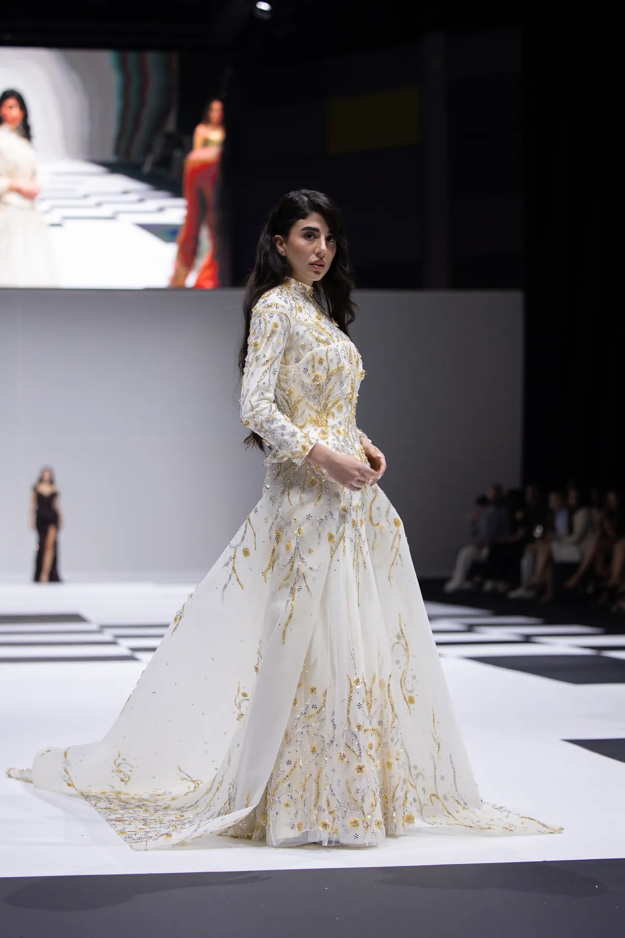 Model walking a fashion runway in a white gown with gold floral embroidery and a long train; dark backdrop and audience visible in the background.