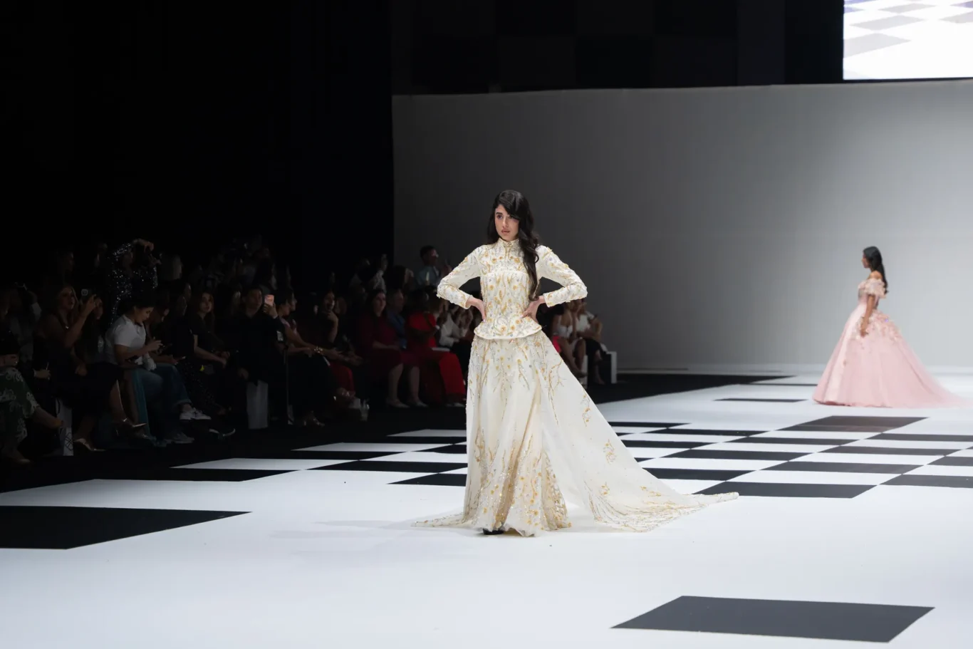 Model in an ivory gown with gold embroidery on a checkerboard runway; audience seated along the left edge of the catwalk.