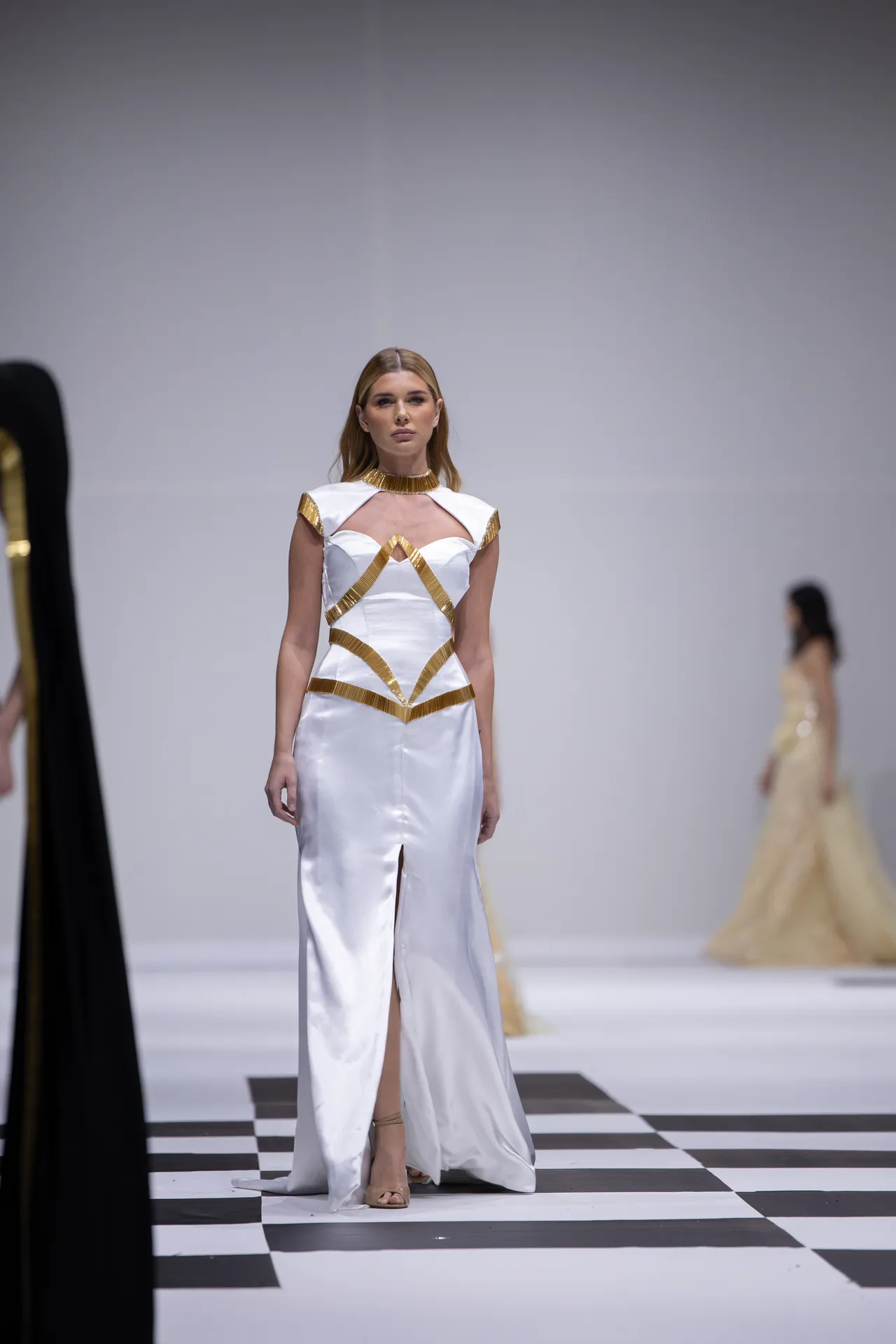 Model walking the runway in a white satin gown with gold geometric straps on a black-and-white checkerboard runway surface.