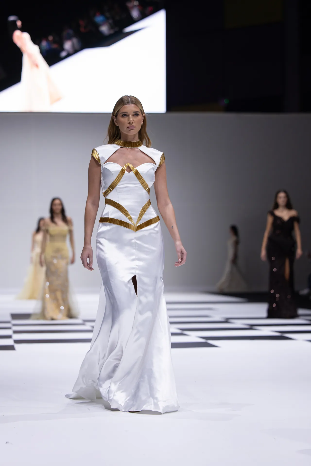 Model walking the runway in a white satin gown with gold geometric straps during a fashion show.