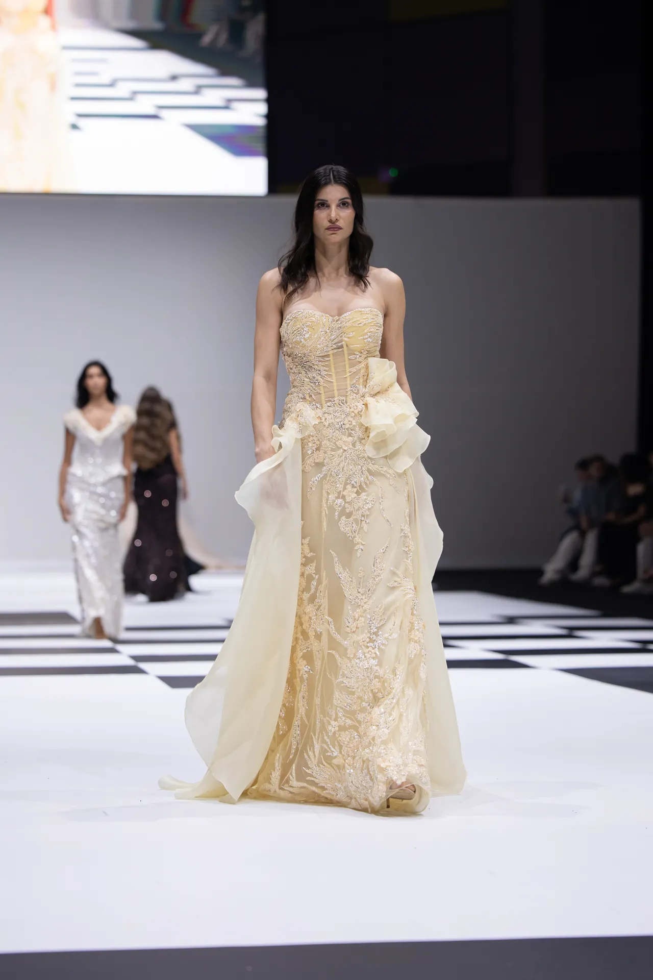 Model walks a fashion runway in a cream, lace-embellished gown with a large ruffle detail on the side; other models visible in the blurred background.