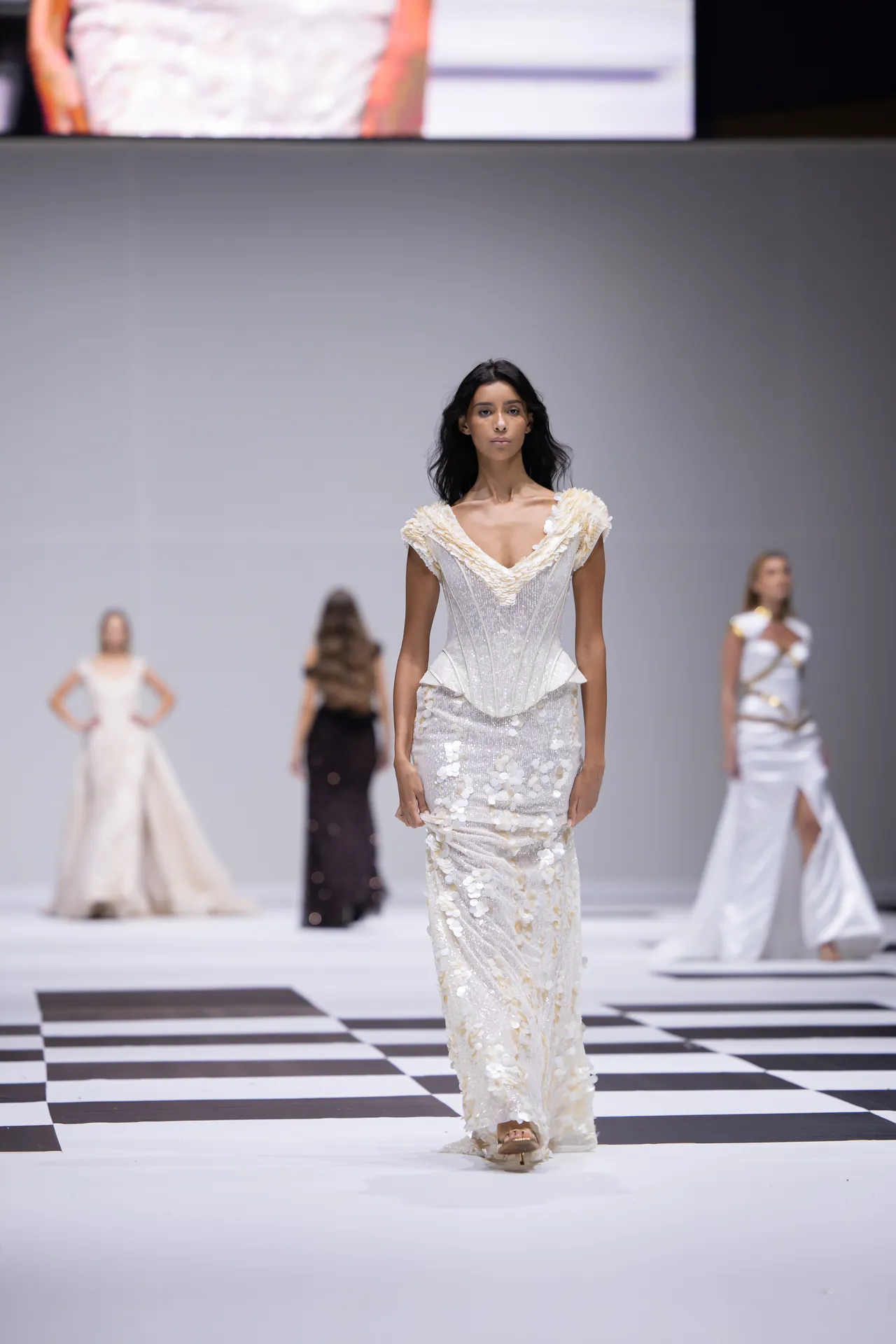 Model walking a runway in a white, beaded gown with a deep V neckline, on a black-and-white checkered catwalk.