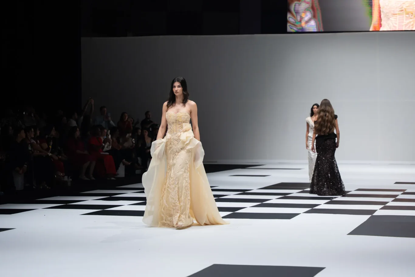Model in a strapless cream-beige gown walks the runway on a black-and-white checkered catwalk, with audience seated in the shadows on the left and other models in the background.