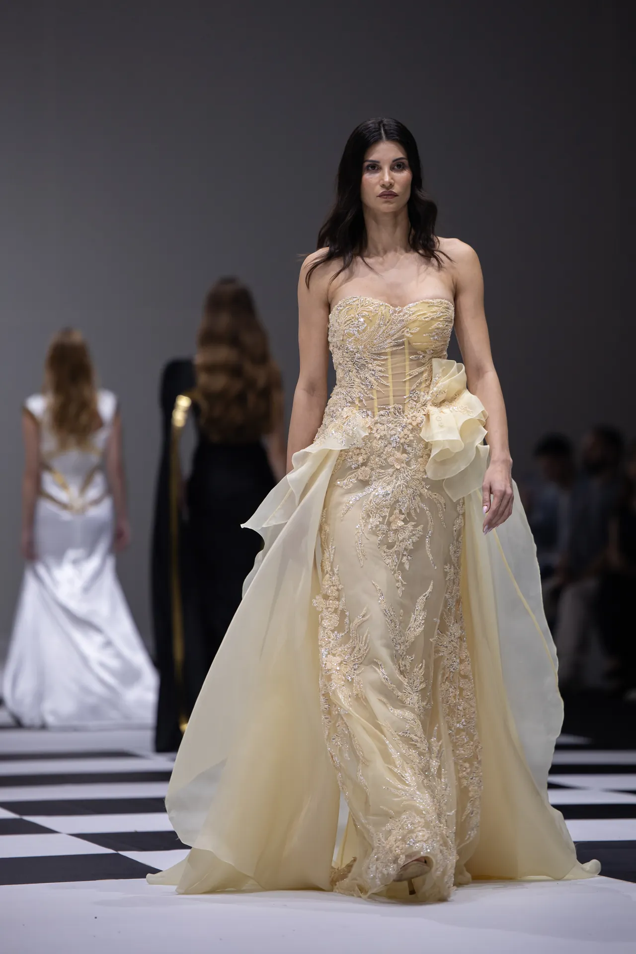 Model walking the runway in an ivory-gold embellished gown with intricate beading and a flowing train