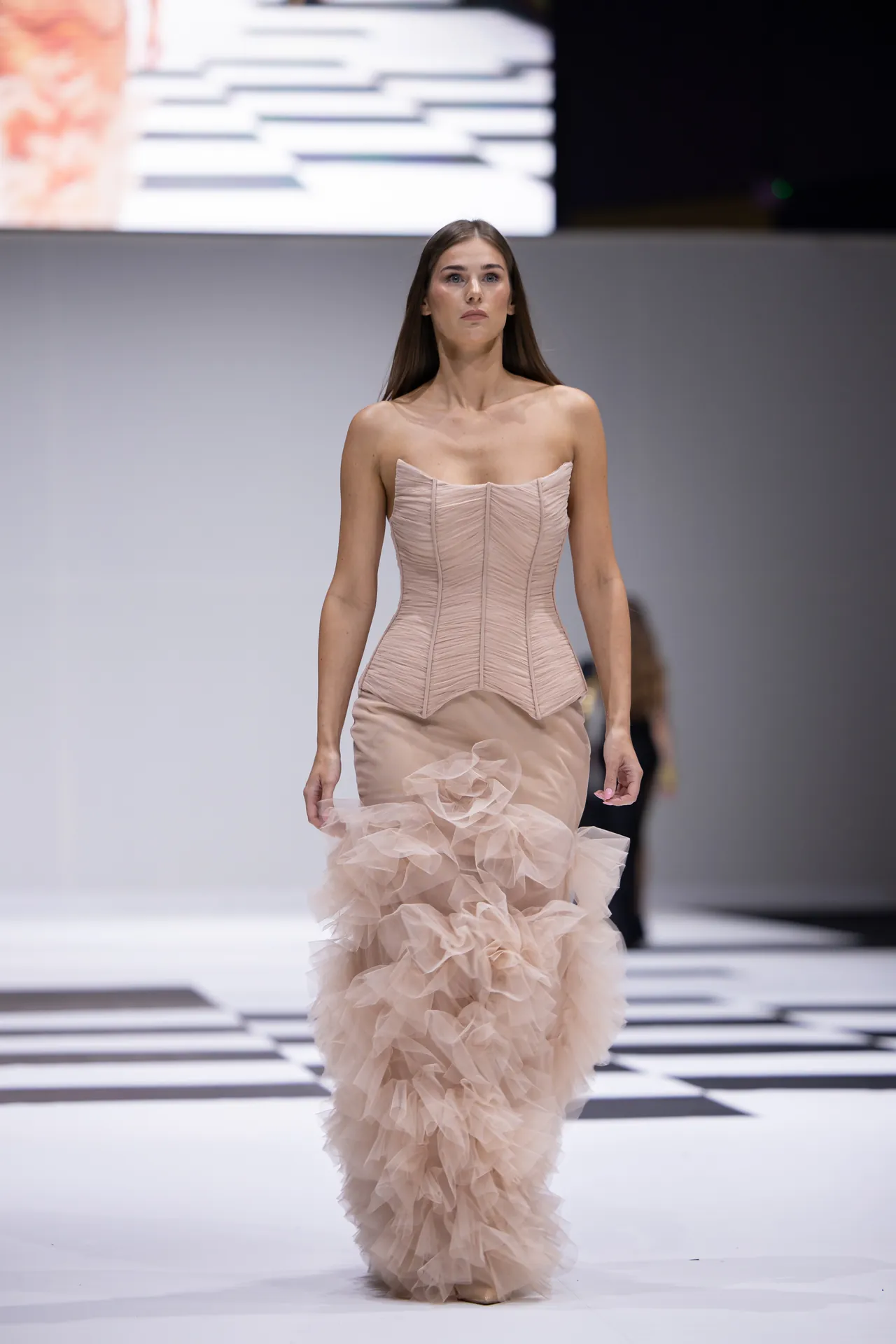 Model walking a fashion show runway in a pale pink strapless corset top with a voluminous layered ruffle skirt.