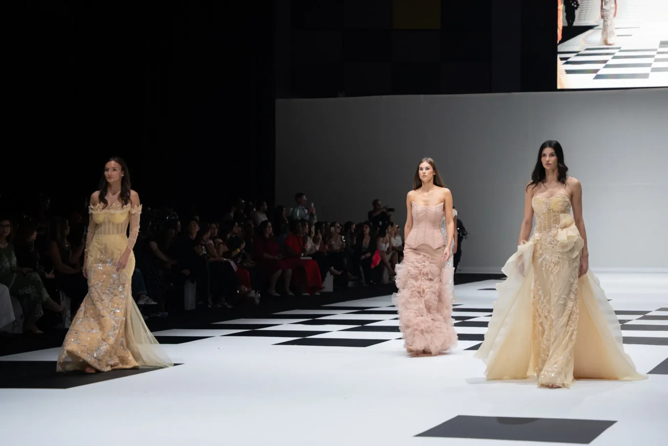 Models in elegant, gown-filled outfits walk a black-and-white checkered runway as the audience watches from the sides.