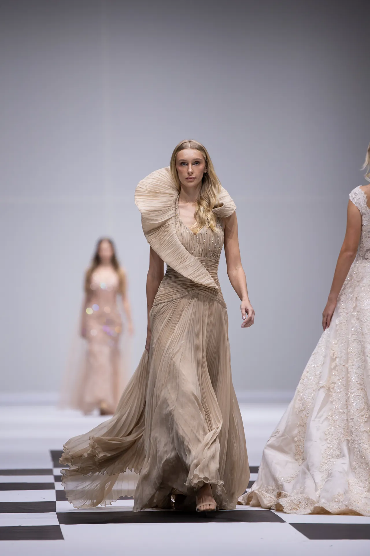 Model walks a runway in a flowing beige gown with a sculptural, oversized shoulder detail on a black-and-white checkered floor.
