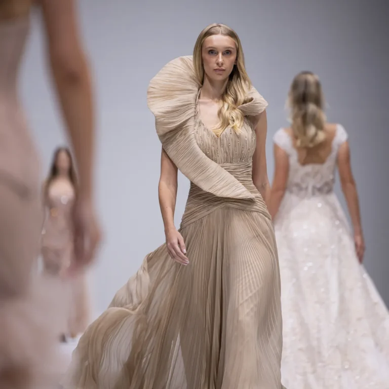 Model walking a beige pleated gown down the runway with other blurred models behind her.