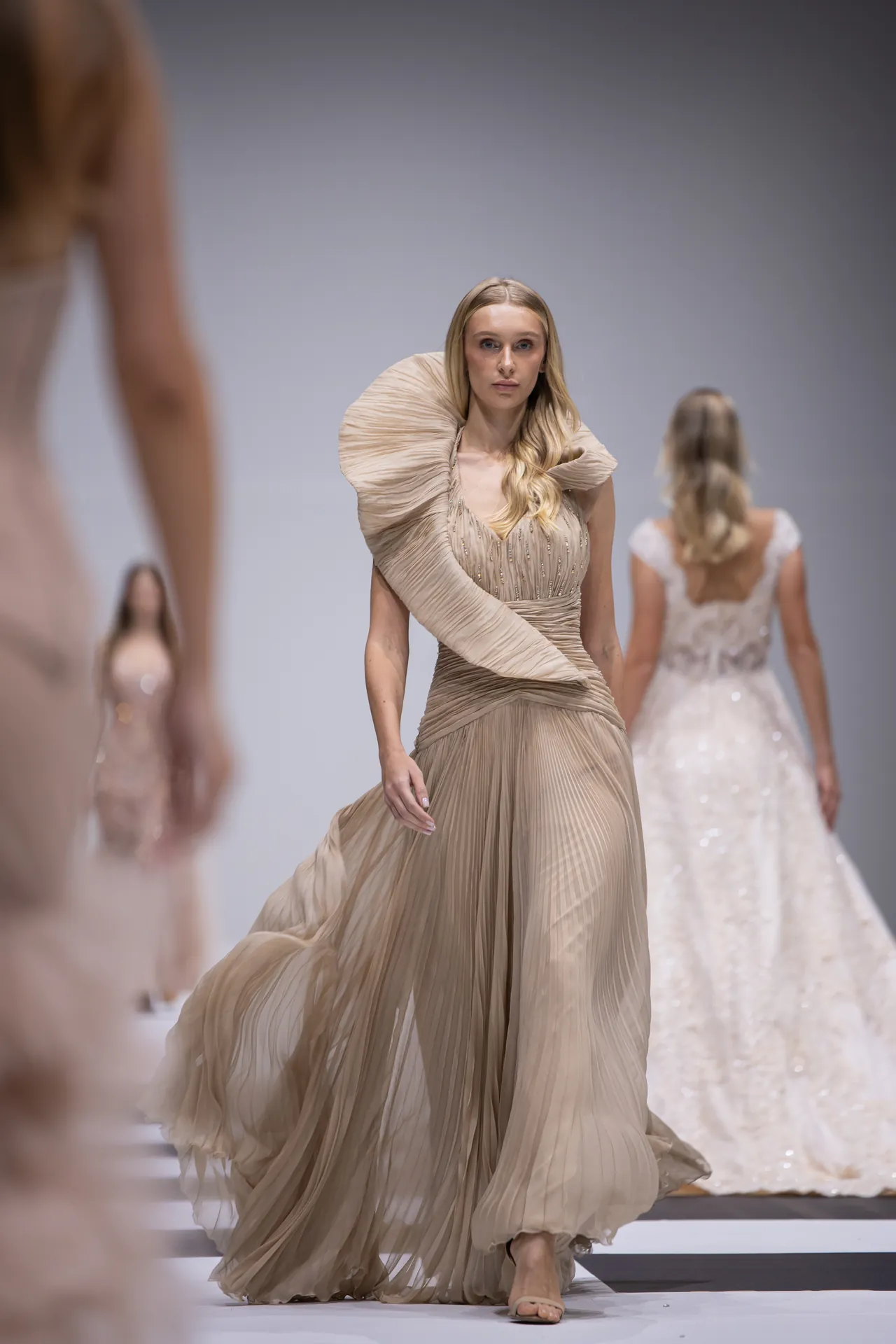Model walking a beige pleated gown down the runway with other blurred models behind her.