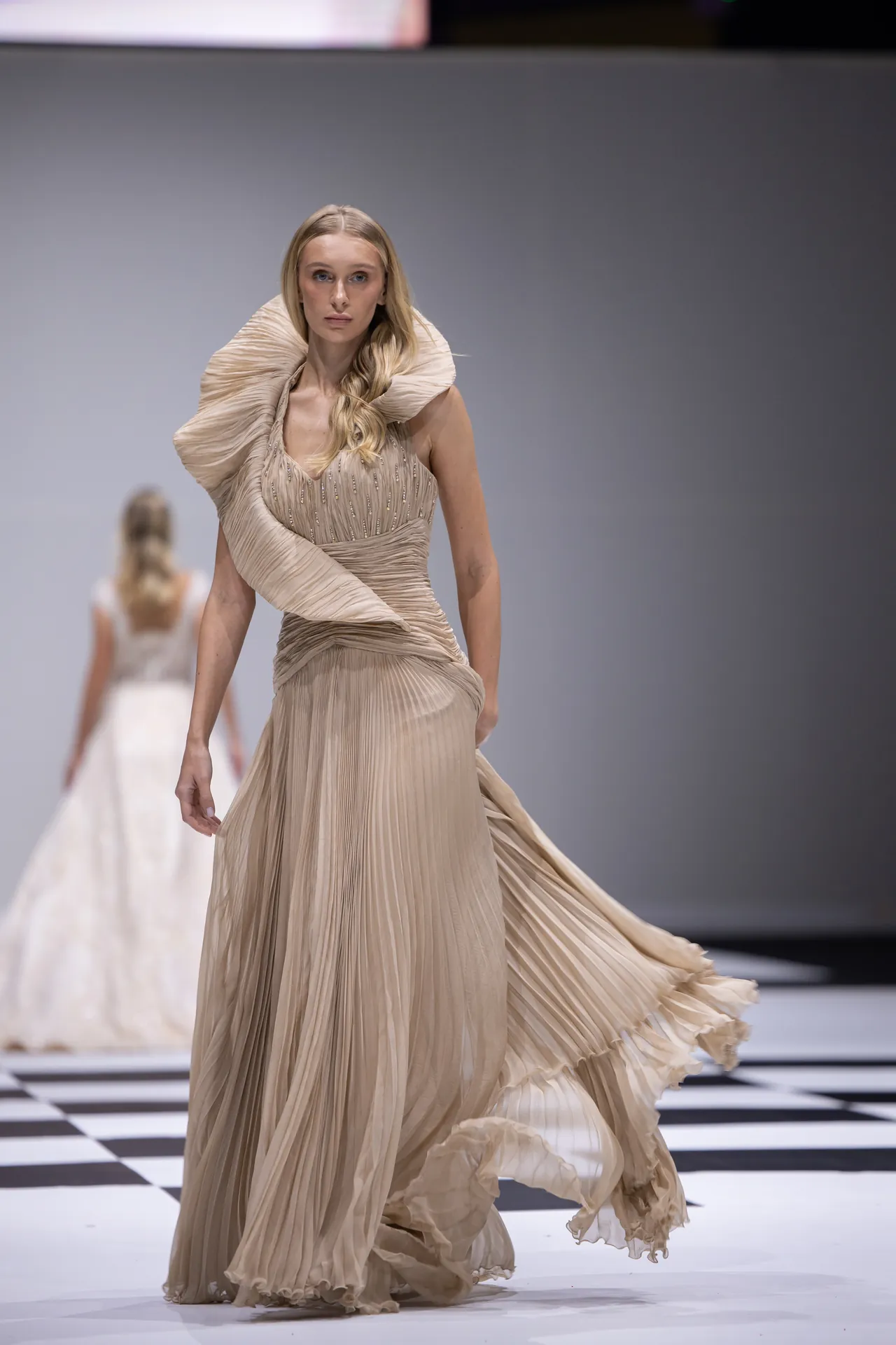 Female model walks a champagne-beige, pleated gown with a large sculptural collar on a black-and-white checkered runway during a fashion show.
