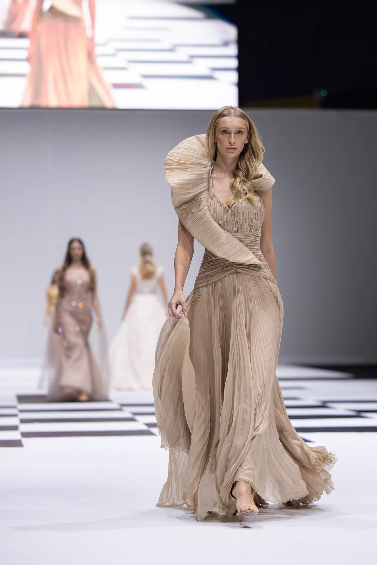 Model walks the runway in a flowing beige gown with dramatic pleated shoulders on a black-and-white checkered catwalk, blurred background models.