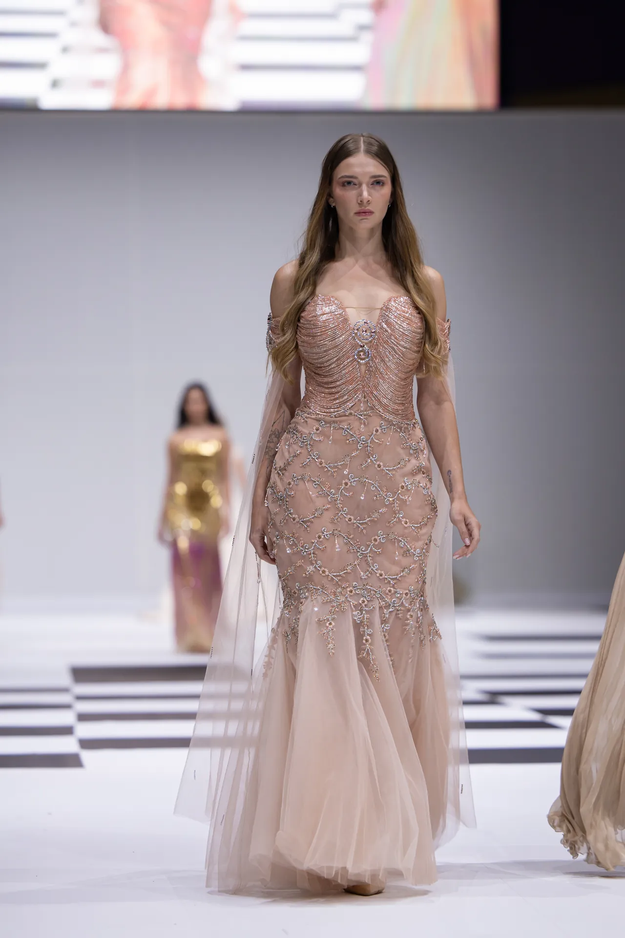 Runway model in a blush pink, floor-length gown with intricate silver embroidery and sheer cape-like overlay.