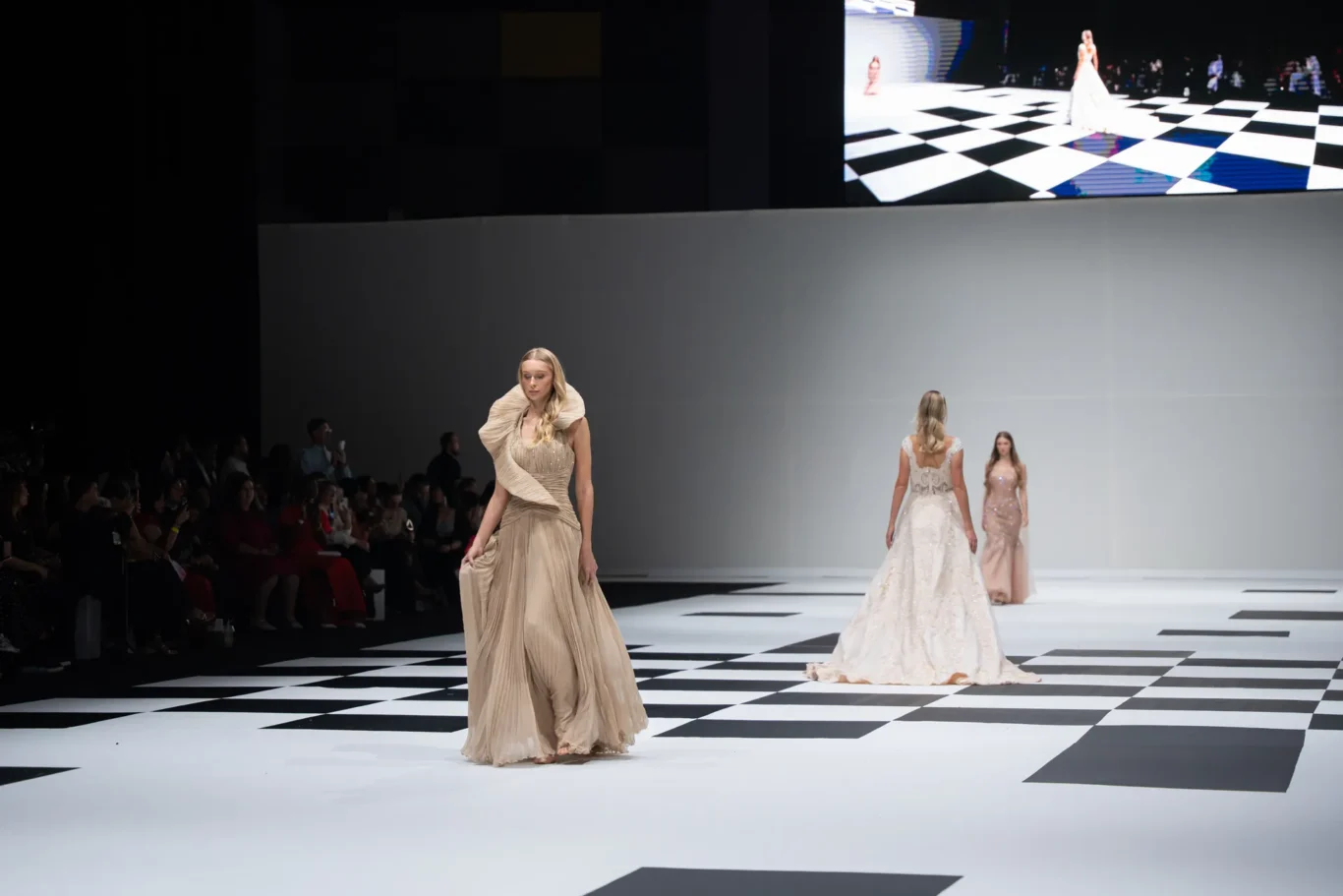 Model in a flowing beige gown walks a black-and-white checkerboard runway; audience seated on the left and a large screen visible above.