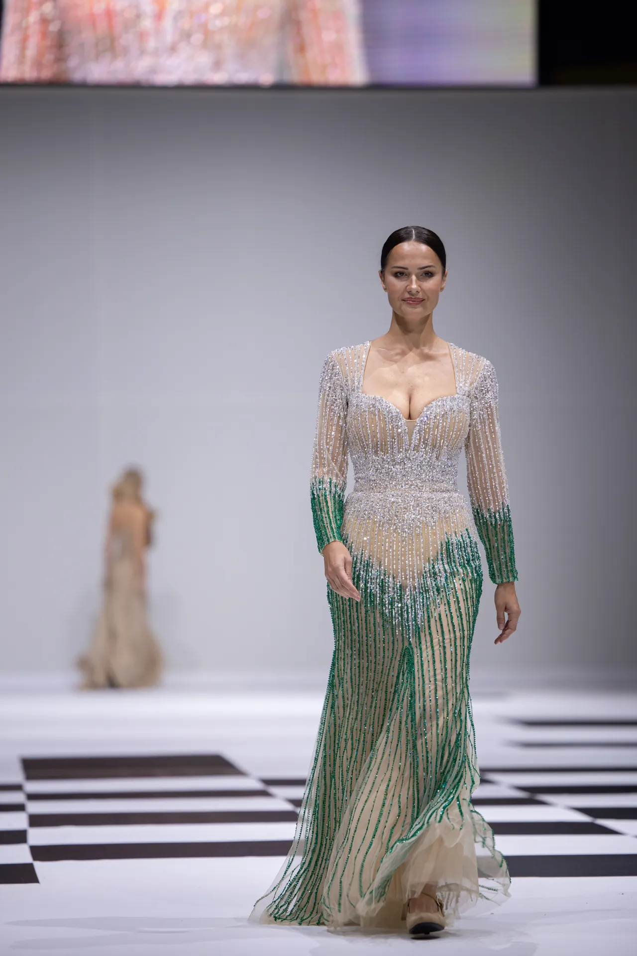 Model walks the runway in a sheer, beaded gown with a green-to-nude gradient and a plunging neckline on a fashion show catwalk.