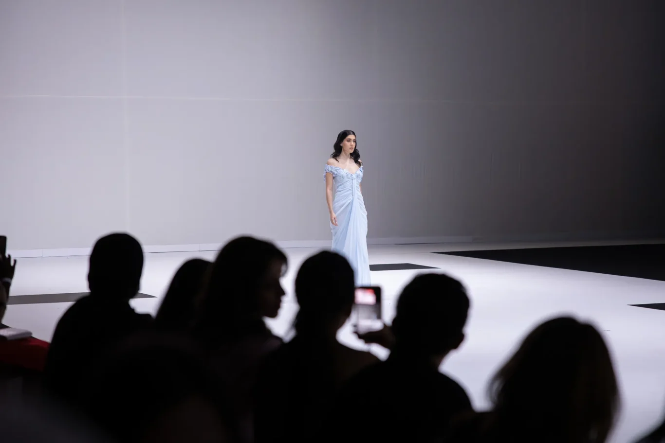 Model in a pale blue off-shoulder gown walking a white runway, audience silhouettes in the foreground.