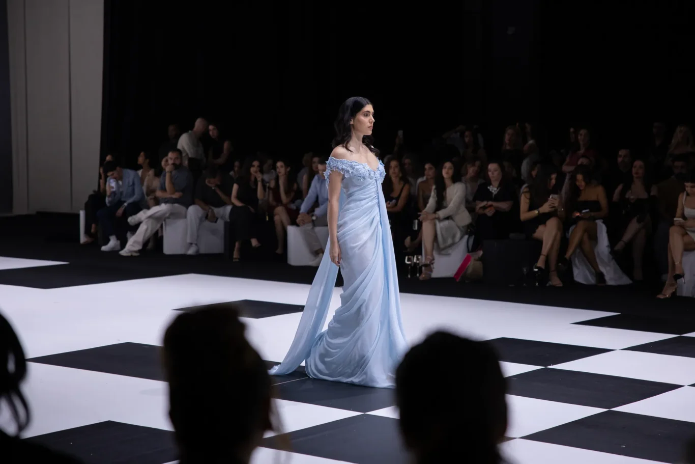 Model in a light blue off-shoulder gown walking a black-and-white checkerboard runway, audience seated behind in dim lighting.