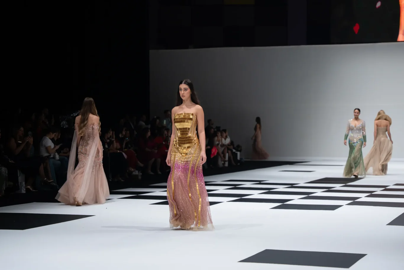 Model walks a fashion runway in a gold and pink sequined gown; black-and-white checkered floor and audience seated along the sides.