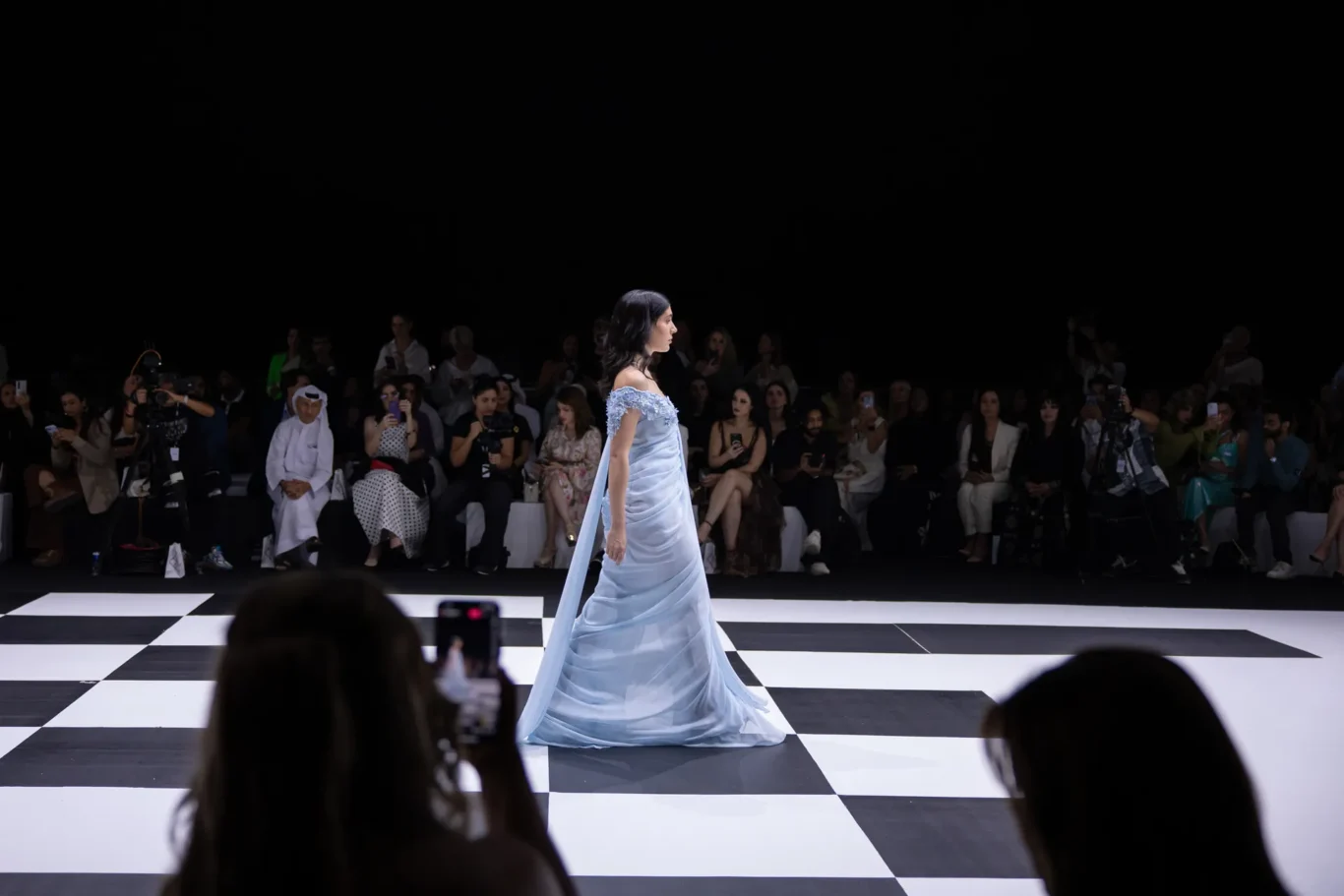 Model in a light blue off‑the‑shoulder gown walks a black-and-white checkered runway as photographers and an audience watch.