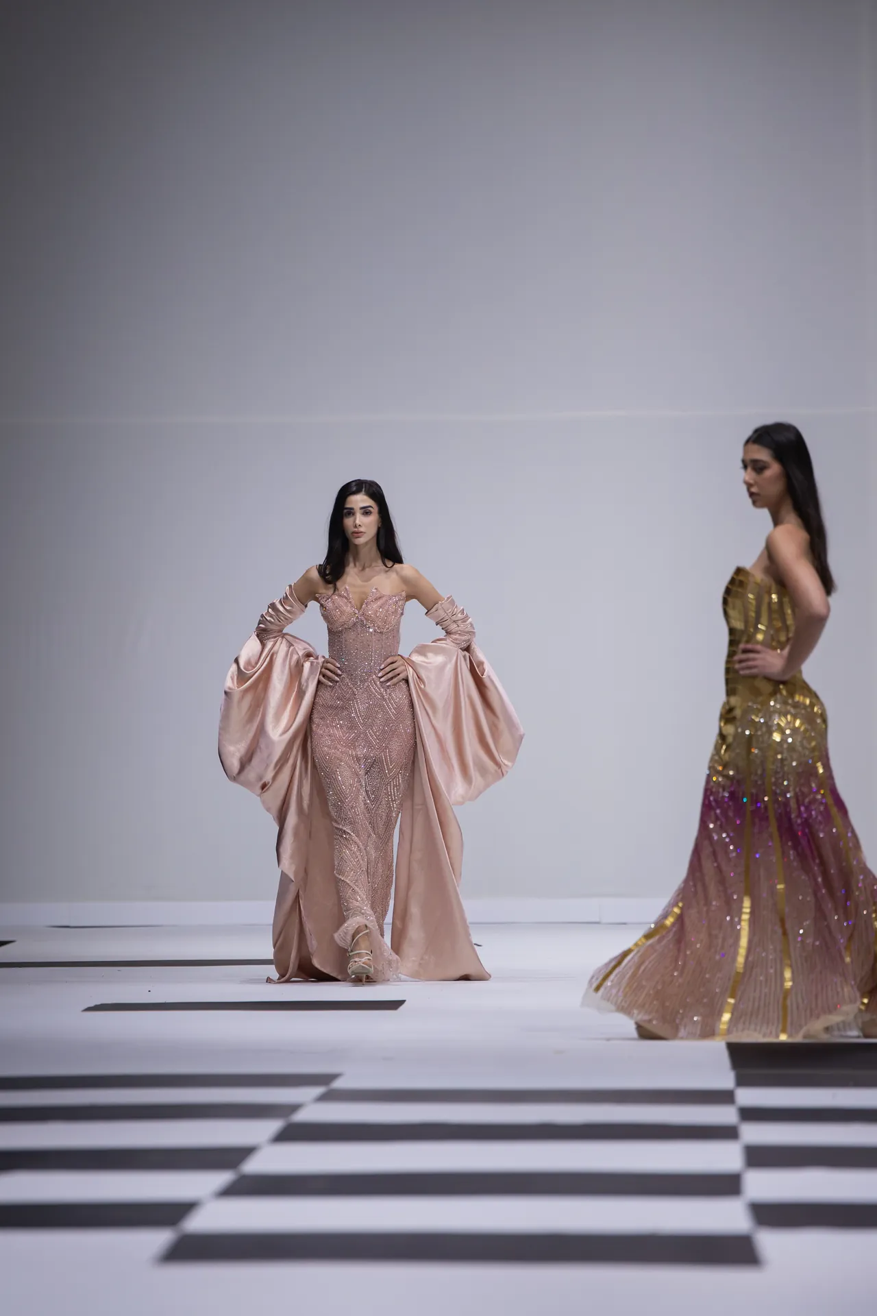 Two models walk a fashion runway in elegant gowns; the foreground model wears a pale pink beaded gown with voluminous satin sleeves, while another in a gold-toned dress stands in the background on a black-and-white checkered floor.
