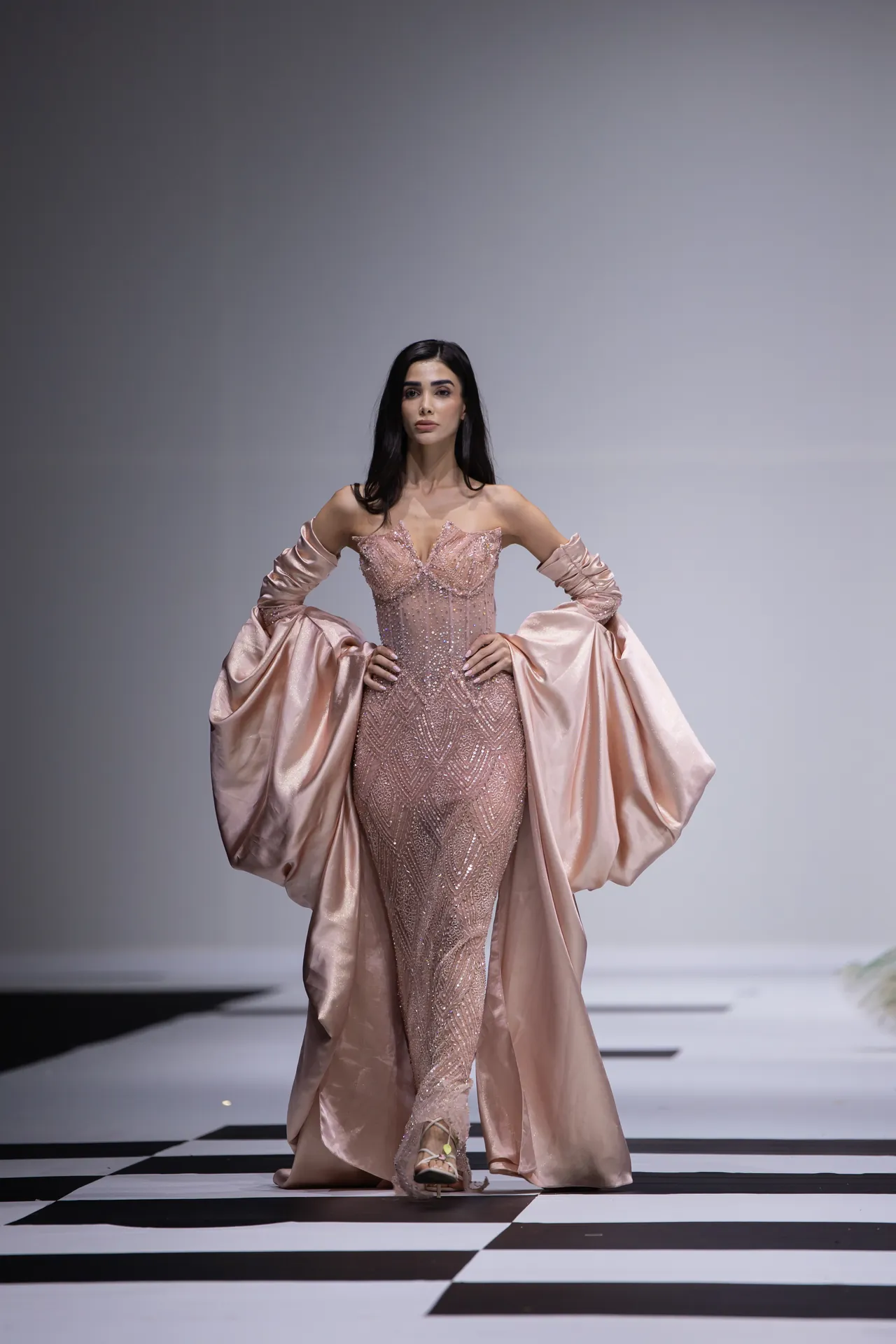 Model walking the runway in a shimmery pink gown with flowing satin cape sleeves.