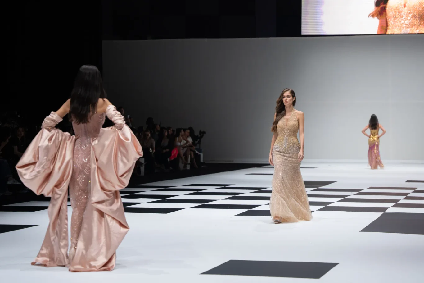 Model in a glittering champagne gown walks the black-and-white checkered runway; pink gowned model appears on the left edge in the background.