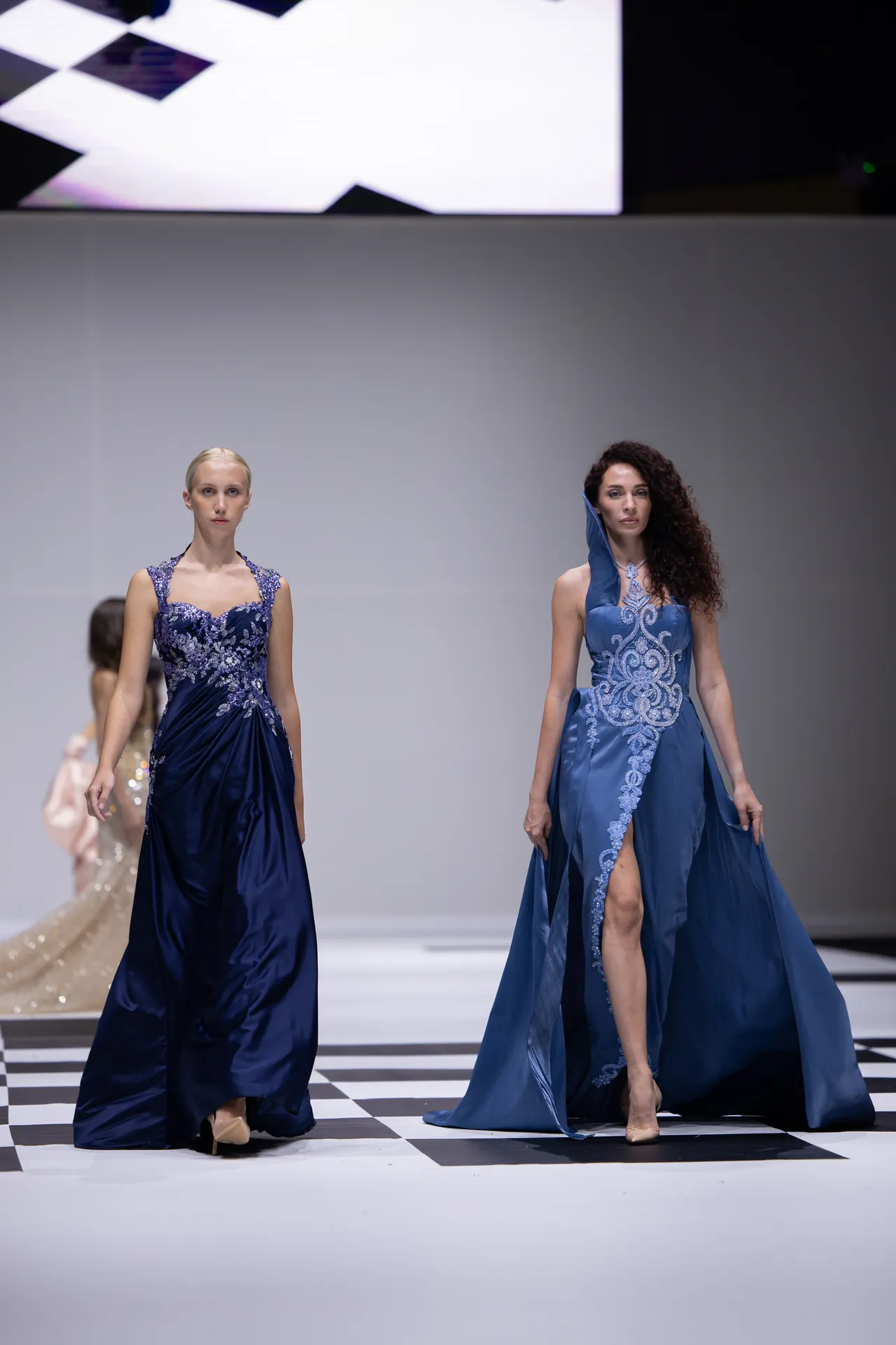 Two models walk a runway in blue gowns with lace embellishments on a black-and-white checker floor.