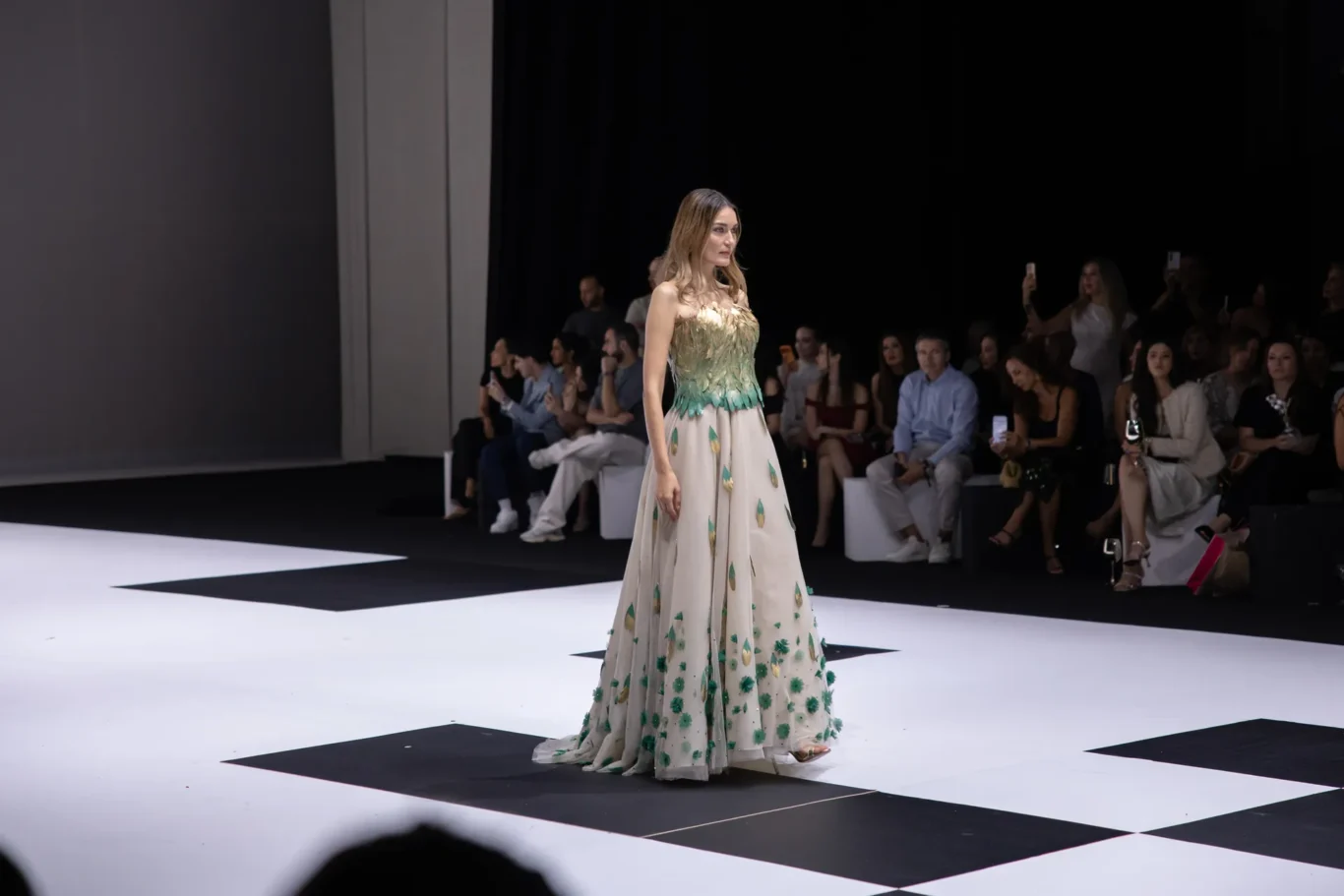 Female model on a white runway wearing a strapless cream gown with green leaf appliqués; audience seated in background taking photos and videos.