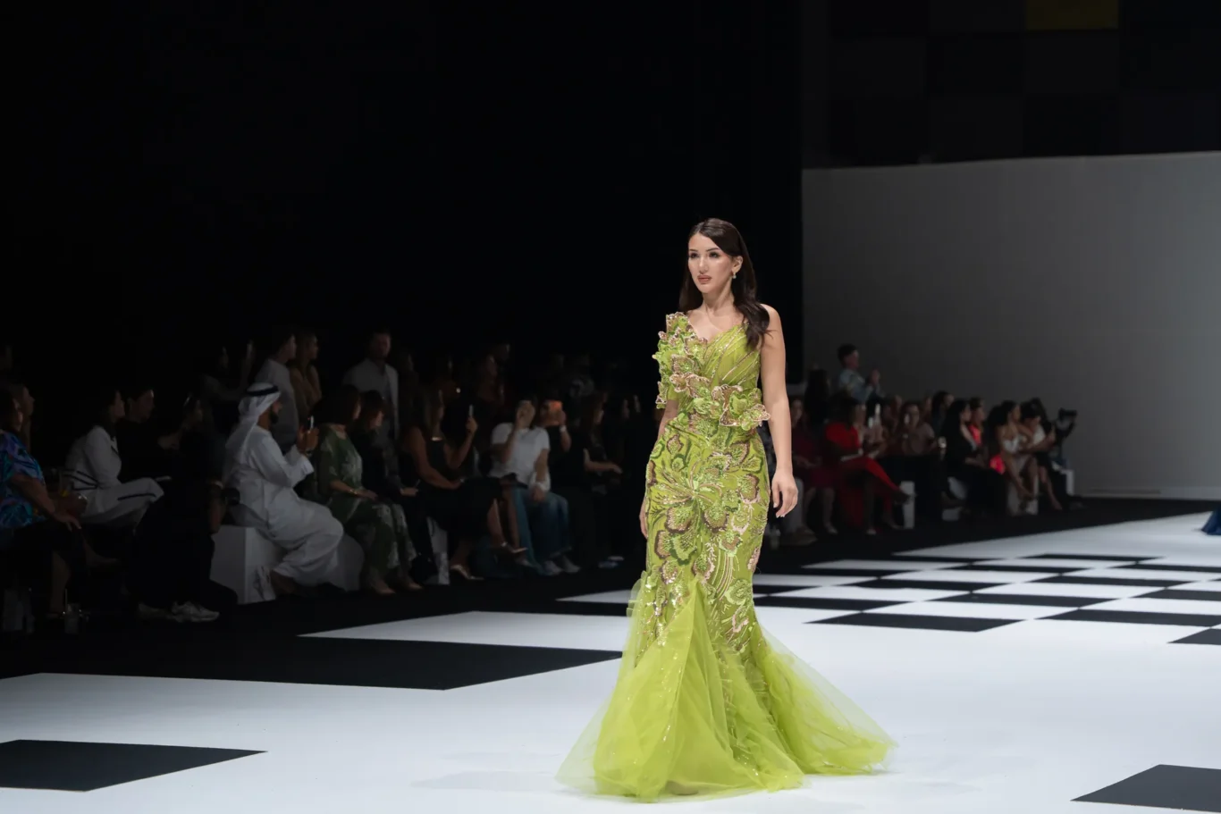 Model walks a lime-green, heavily embroidered gown down a fashion runway; audience seated along dark sidelines gaze on.