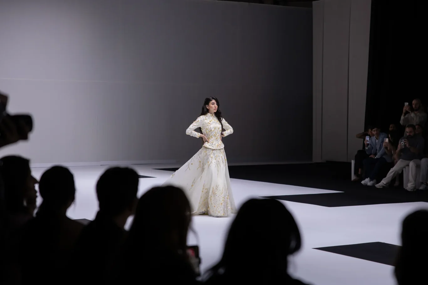 Model on a white runway wearing a white and gold-embroidered gown, audience on the right filming with their phones.
