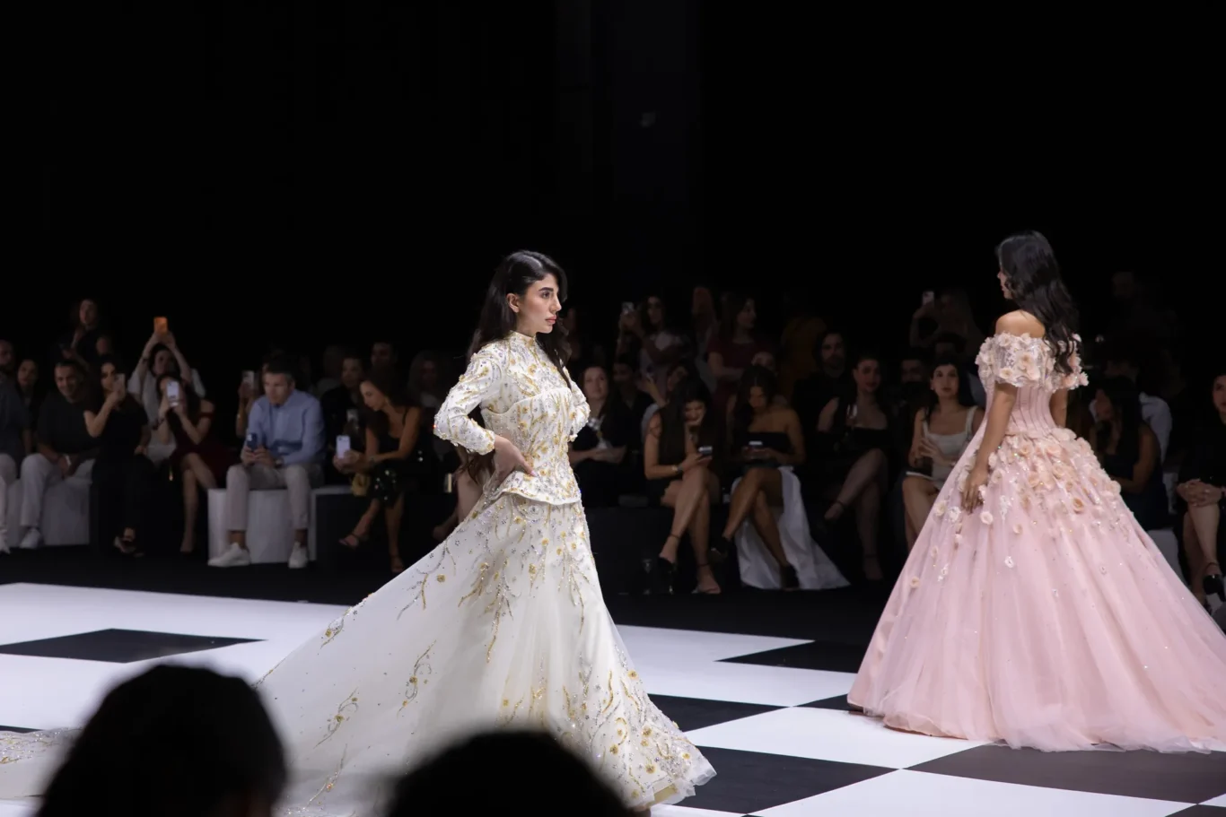 Two models in elaborate gowns walk a fashion runway as an audience watches and photographs.