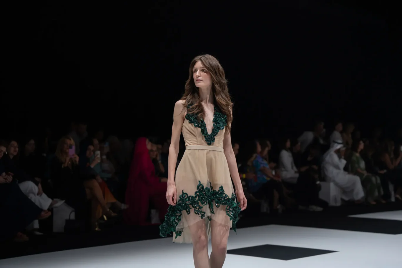 Model walking the runway in a beige dress with dark green floral embroidery; audience blurred in the background.