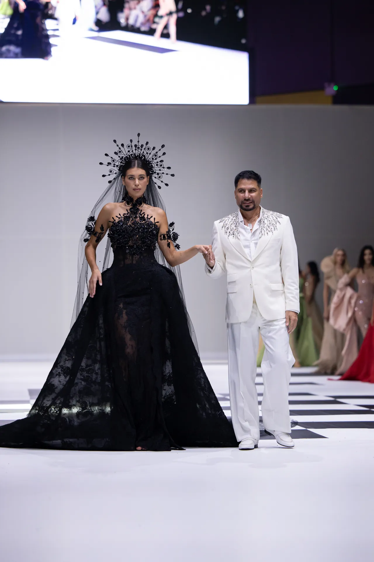 Female model in a black lace ball gown with an ornate headpiece walks the runway, holding hands with a man in a white suit with silver embellishments.