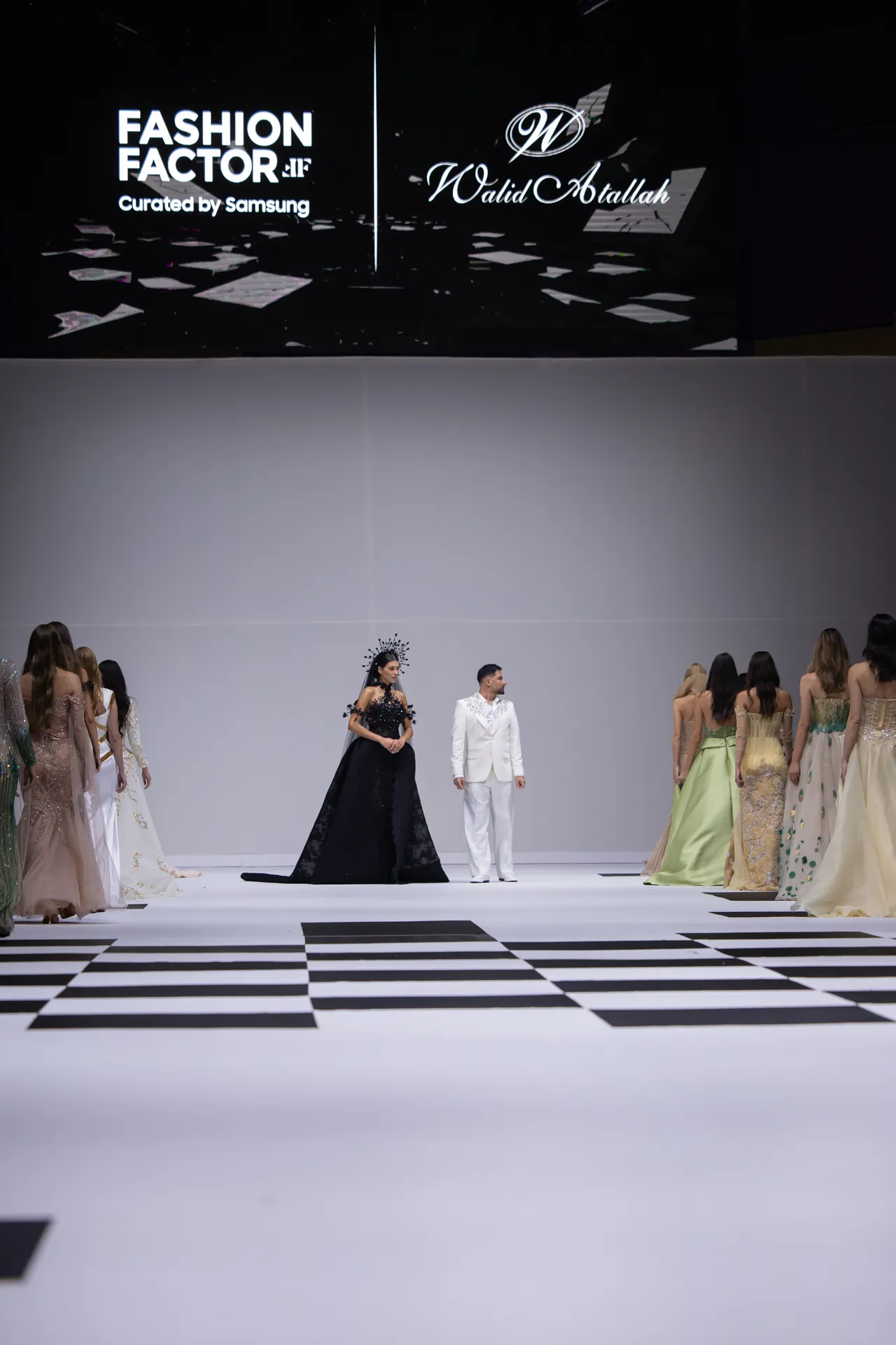 Center-stage fashion show: a woman in a black ball gown and a man in a white suit stand on a white runway with a bold black-and-white checker floor, surrounded by models in pastel gowns; large screen behind reads 'Fashion Factor'.