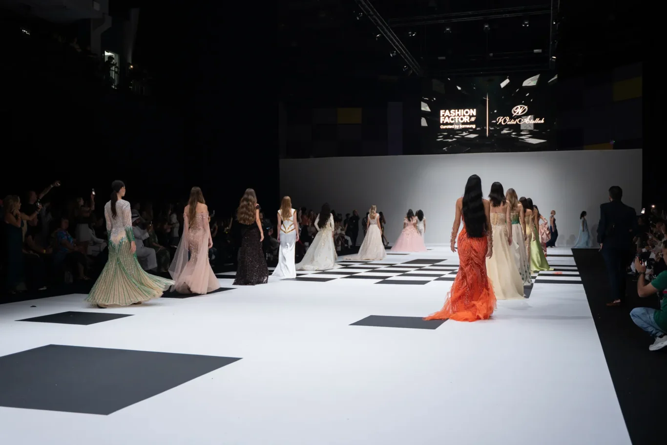 Models wearing elaborate gowns walk down a black-and-white checkerboard runway at a fashion show, audience on both sides, large screen stage in back.