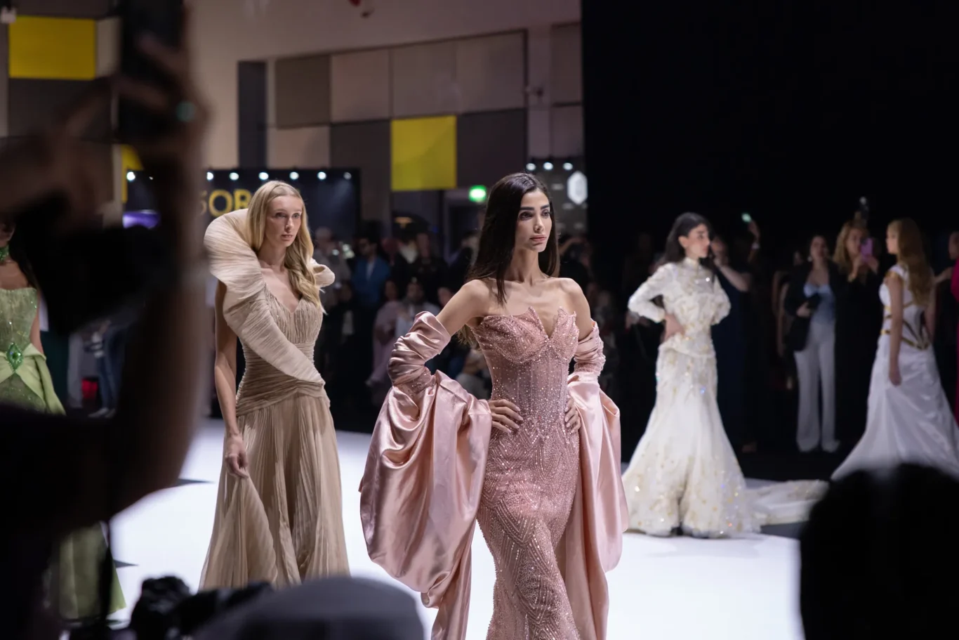 Fashion models wearing glamorous gowns walk the white runway at a crowded fashion show, pink beaded dress in foreground center.