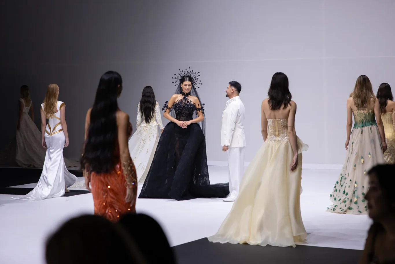 Center-stage woman in an ornate black ball gown with a spiked headpiece on a fashion-show runway, surrounded by other models in formal gowns farther back on stage, audience silhouettes visible in foreground.