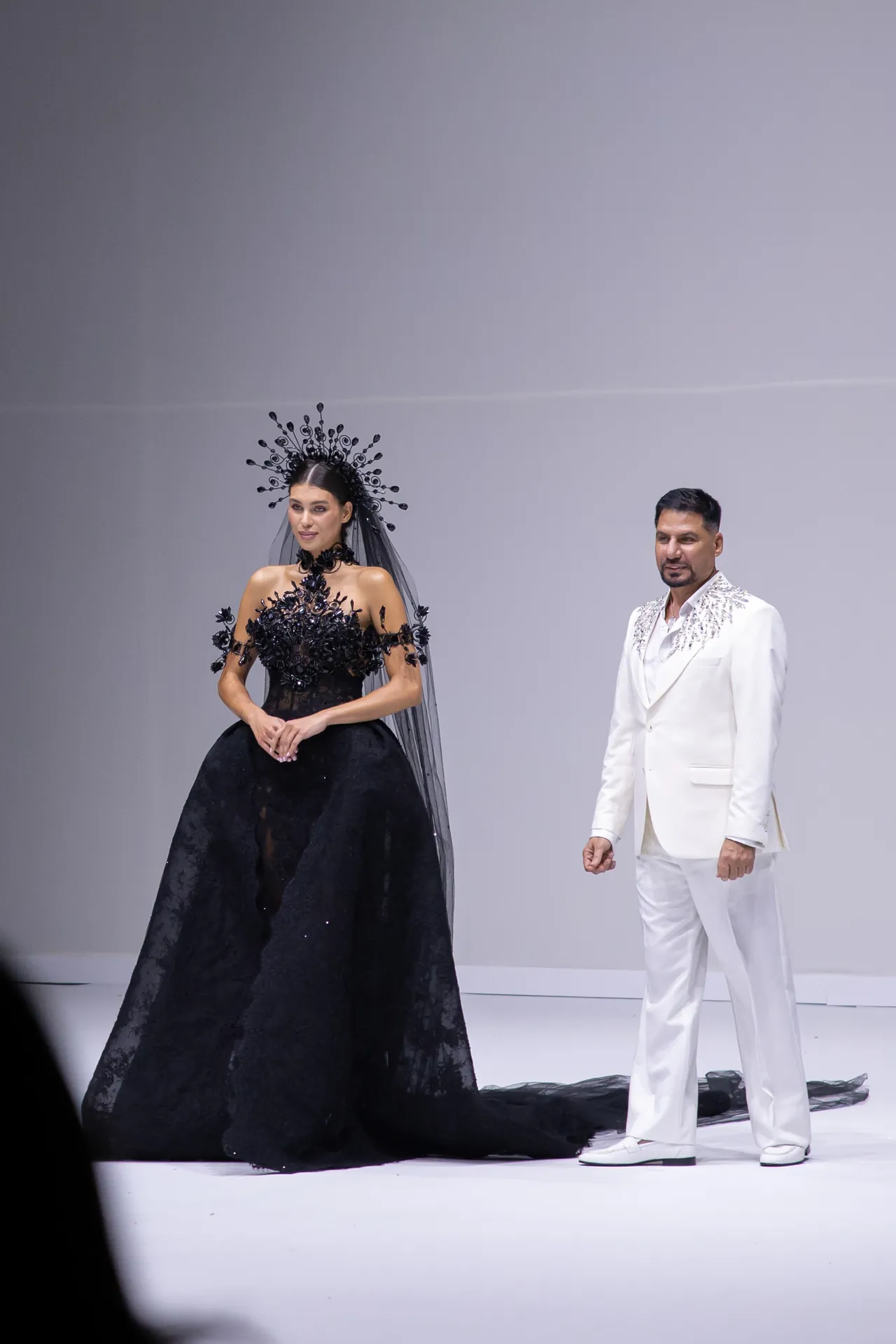 Woman in an elaborate black ball gown with floral appliqués and a dramatic headpiece stands on a catwalk beside a man in a white embroidered suit. Women’s fashion show scene, studio backdrop.