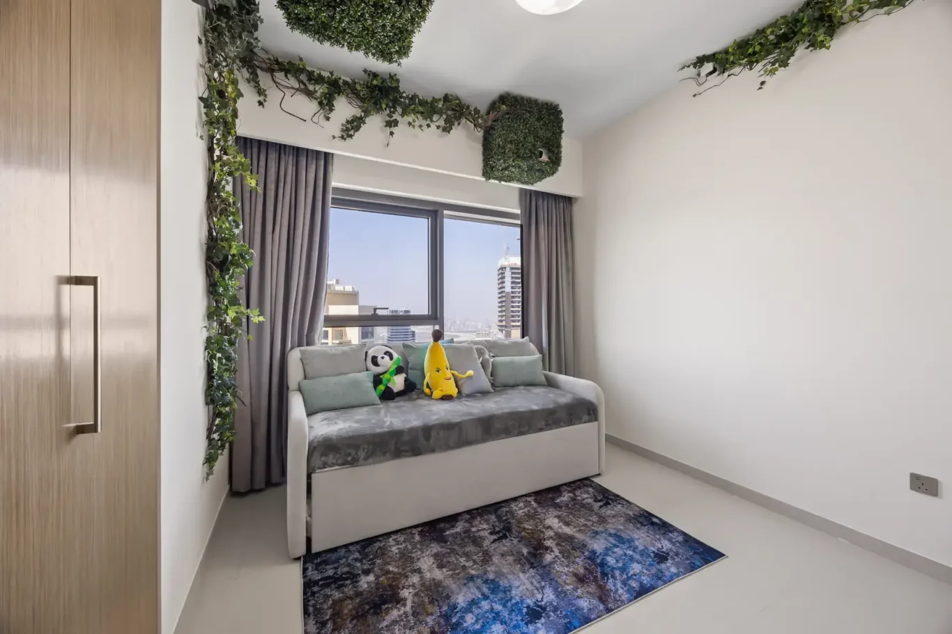Bright small living room with a gray sofa under a city-view window, green ivy decor along the ceiling, and panda and banana plush toys on cushions and throw pillows.