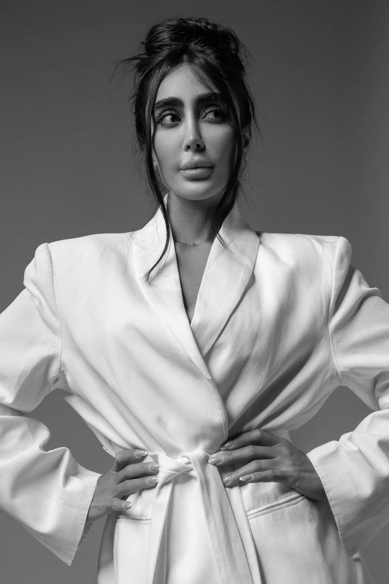 Woman in a white blazer tying the belt, looking off to the side in a black-and-white studio portrait.