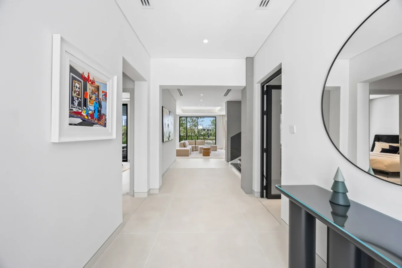 Bright white hallway leading to a living room with large windows; framed art on the left and a round mirror on the right.
