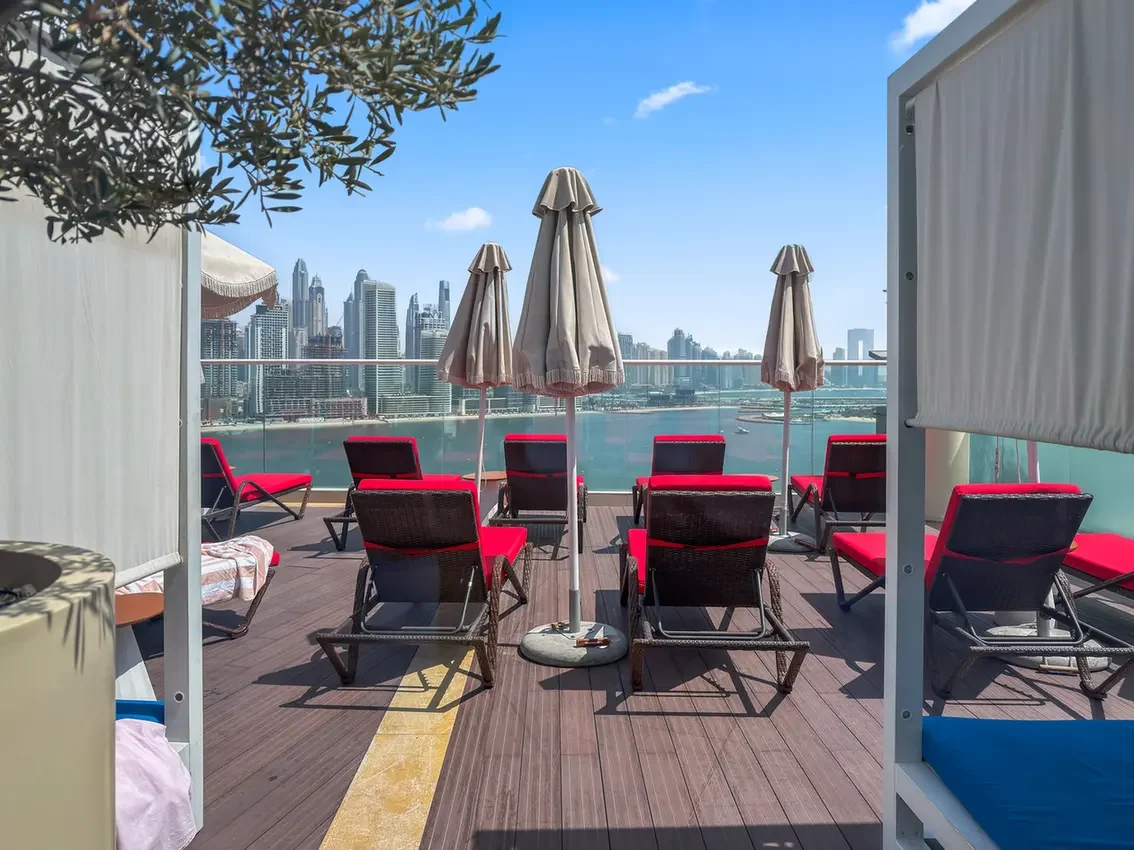 Rooftop pool deck with red cushioned lounge chairs and beige umbrellas, overlooking a city skyline by the water.