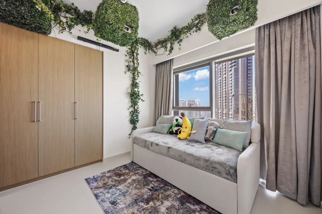 Modern bedroom with a white and grey sofa by a large window; wooden wardrobe on the left and ivy creeping along the ceiling above.