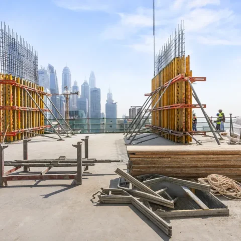 Construction site on a rooftop with wooden and metal formwork, rebar, and workers; city skyline in the distance.